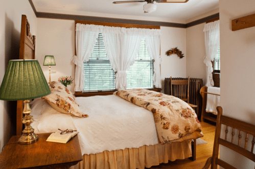 Cozy bedroom with a floral bedspread, green lamp, and two windows dressed with white curtains. Warm, inviting atmosphere.