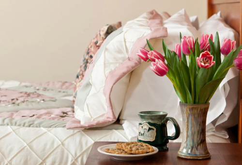 A cozy bedroom scene featuring a quilted bed, a vase of pink tulips, a mug, and a plate of cookies on a bedside table.