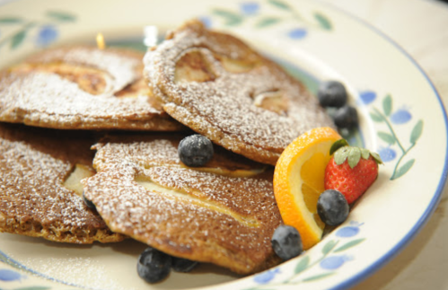 A plate of fluffy pancakes dusted with powdered sugar, garnished with strawberries, blueberries, and orange slices.