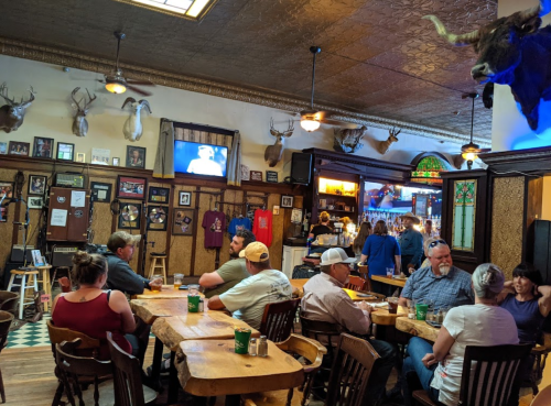 A lively bar scene with patrons seated at wooden tables, deer heads on the walls, and a TV in the background.