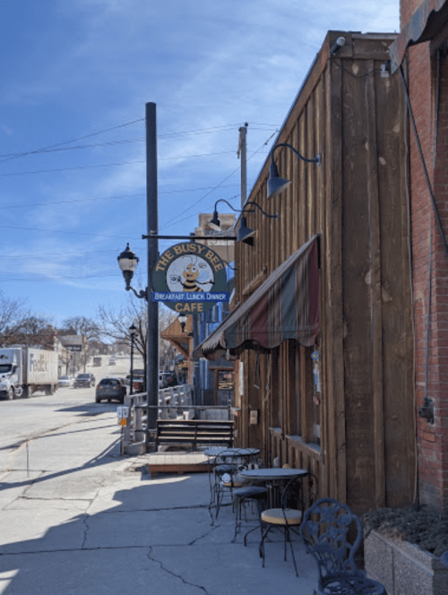 A street view of a café called "The Busy Bee," featuring outdoor seating and a clear blue sky.