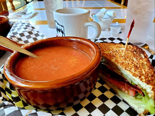 A bowl of tomato soup next to a grilled cheese sandwich with lettuce and tomato, served on a checkered plate.