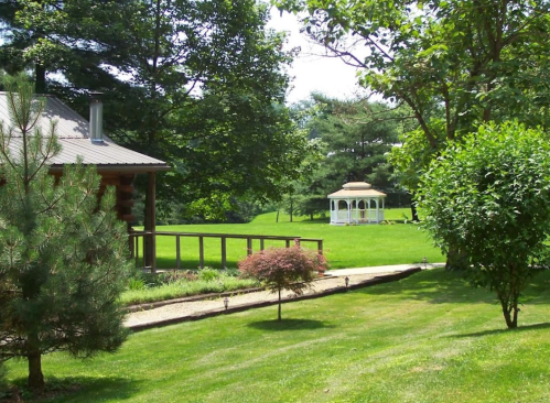 A serene landscape featuring a gazebo, lush green grass, trees, and a log cabin in the background.