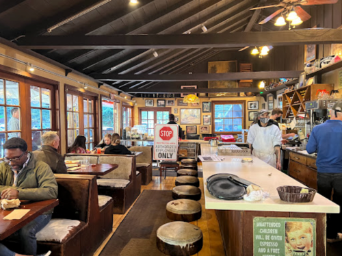 Cozy café interior with wooden beams, patrons seated at tables, and a counter with a barista serving coffee.