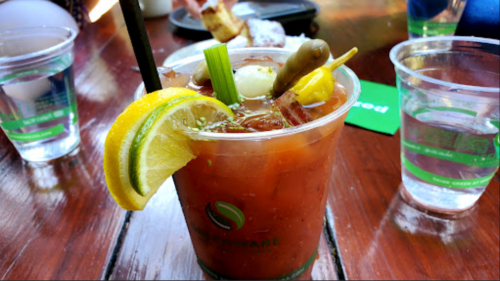 A refreshing cocktail in a clear cup, garnished with lemon, celery, olives, and a pickle, on a wooden table.