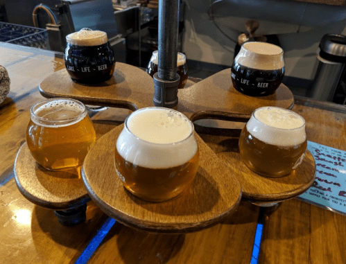 A wooden flight holder with six small glasses of different beers, including light and dark varieties, on a bar table.