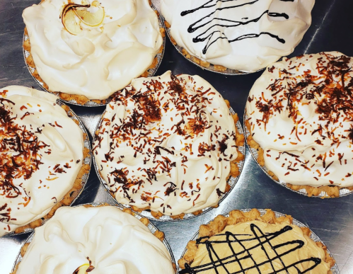 A variety of cream pies topped with whipped cream, chocolate drizzle, and coconut flakes, arranged on a silver surface.