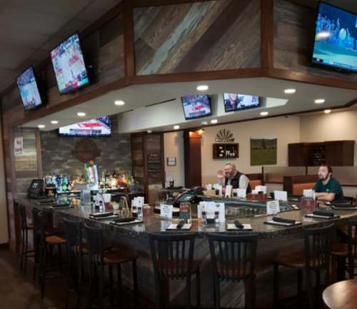 A cozy bar area with multiple TVs showing sports, featuring two patrons and a well-stocked counter.