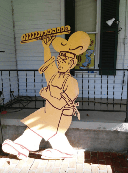 A wooden cutout of a chef holding a long loaf of bread, standing on a brick walkway in front of a house.