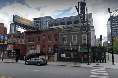 A street view of a historic building with a green facade, adjacent to modern buildings and a billboard.