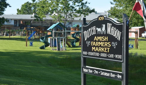 Sign for Dutch Wagon Amish Farmers Market with playground equipment in the background, surrounded by green grass.