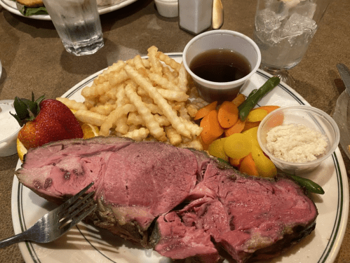 A plate featuring sliced roast beef, crinkle-cut fries, mixed vegetables, a strawberry, and dipping sauce.