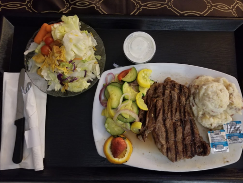 A tray with a grilled steak, mashed potatoes, mixed salad, and a peach, accompanied by ranch dressing and utensils.