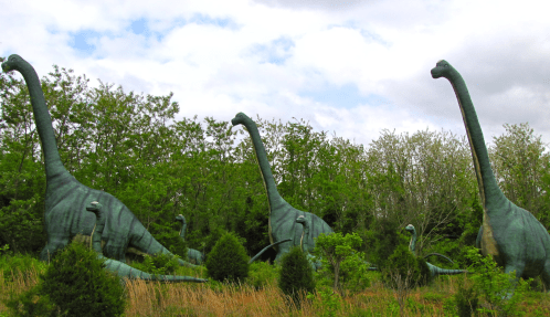 A group of large dinosaur sculptures surrounded by greenery under a cloudy sky.