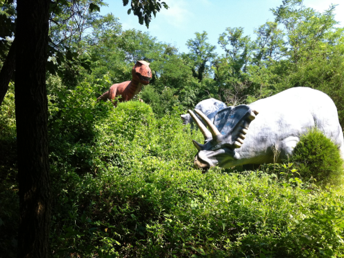 Two large dinosaur sculptures, a T-Rex and a triceratops, partially hidden in lush greenery under a blue sky.