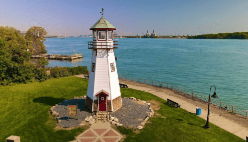 A white lighthouse stands by a calm blue river, surrounded by green grass and benches along the waterfront.
