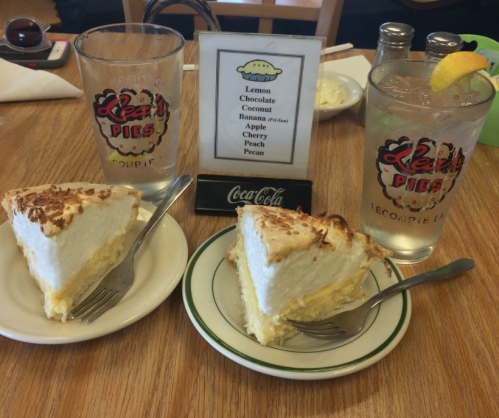 Two slices of pie with whipped cream on plates, alongside a glass of water with a lemon slice and a menu of pie flavors.