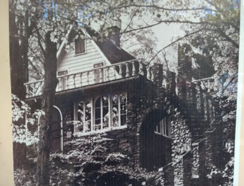 A vintage black-and-white photo of a stone house surrounded by trees, featuring a balcony and intricate architecture.