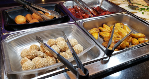 A buffet display featuring sesame balls, sweet plantains, and various meats in metal trays with serving tongs.