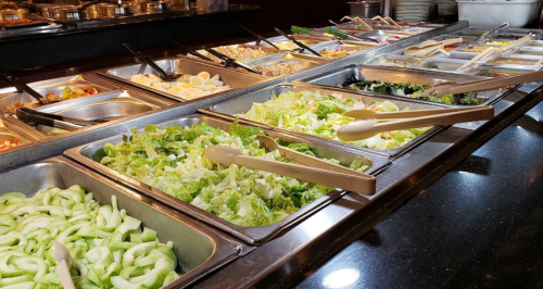 A buffet spread featuring various fresh salads and toppings in metal trays with serving utensils.