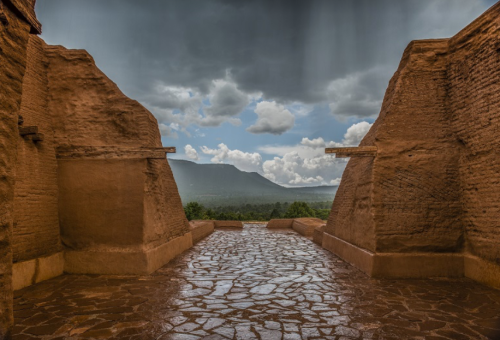 A stone pathway leads to a scenic view of mountains under a cloudy sky, framed by ancient walls.