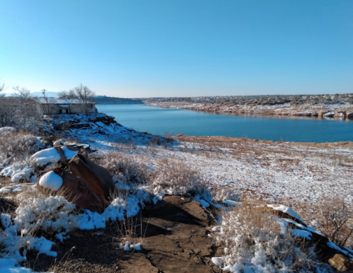 A snowy landscape by a calm lake, with rocky terrain and sparse vegetation under a clear blue sky.