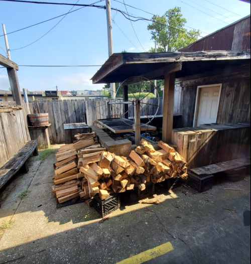 A rustic outdoor space with stacked firewood, wooden benches, and a grill area surrounded by wooden fences.