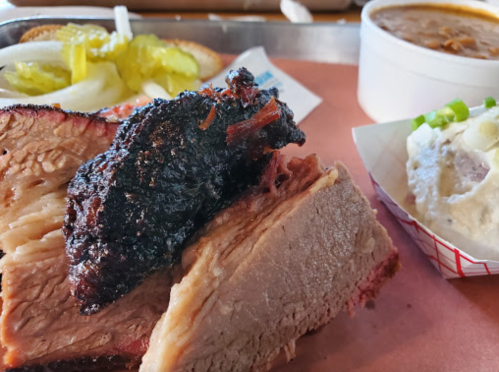 A plate featuring smoked brisket, pickles, onions, bread, and a side of beans and potato salad.