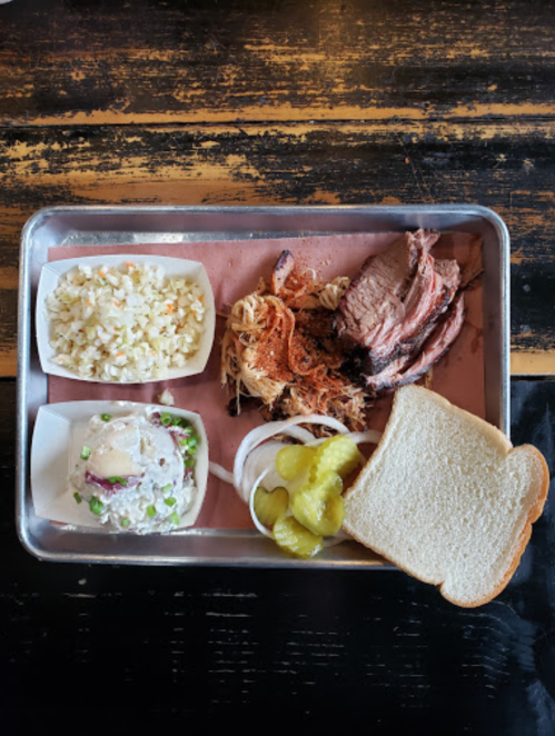 A metal tray with sliced brisket, coleslaw, potato salad, pickles, onions, and a slice of white bread.