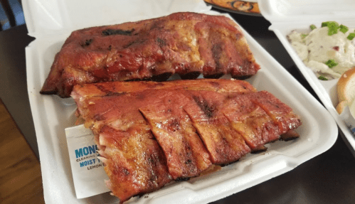 Two racks of glazed, barbecued ribs in a takeout container, with a side of coleslaw visible in the background.
