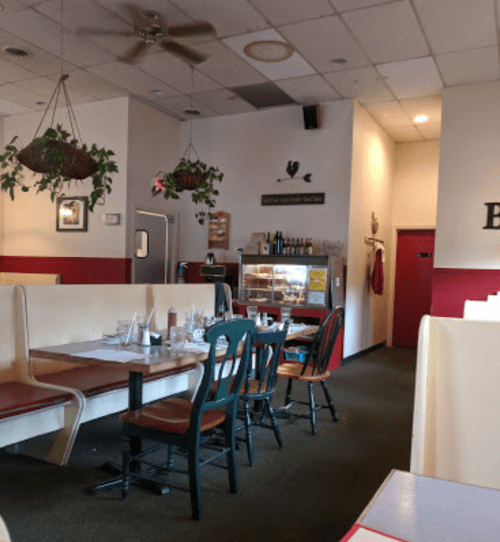 Interior of a casual restaurant with booths, tables, hanging plants, and a display case in the background.