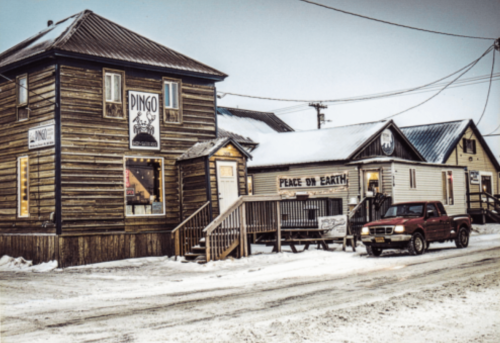 A snowy street scene featuring wooden buildings, including a shop named "Dingo" and a sign that reads "Peace on Earth."