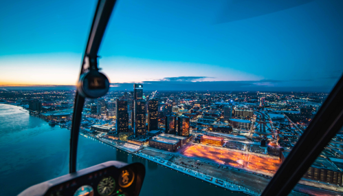 Aerial view of a city skyline at dusk, with lights reflecting on the water and a helicopter cockpit in the foreground.