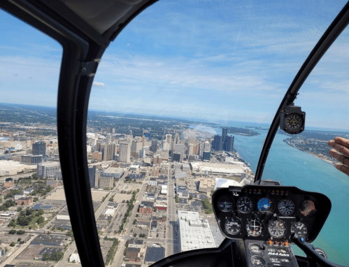 View from a helicopter cockpit overlooking a city skyline and a river on a clear day.