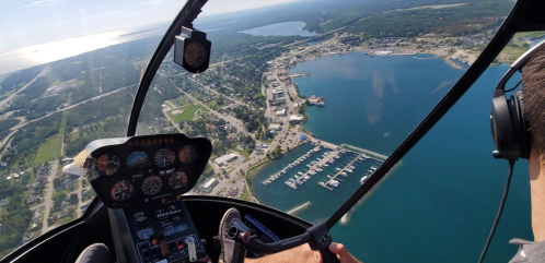 Aerial view from a helicopter showing a marina, town, and water, with a pilot's hands on the controls.