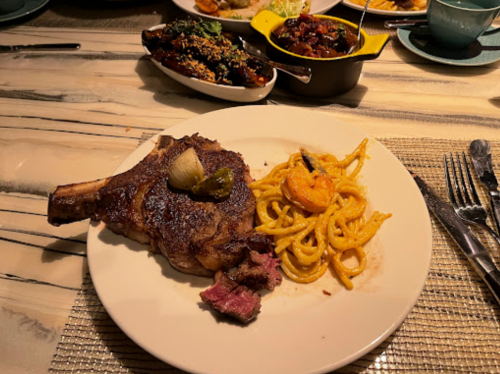 A plate featuring a large steak with an onion garnish, served alongside shrimp pasta on a textured table setting.