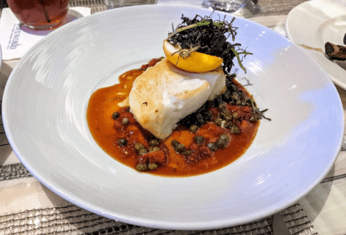 A white fish fillet topped with greens and lemon, served on a bed of tomato and caper sauce in a white bowl.