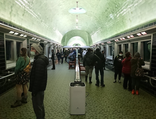 A subway station with a green-tiled archway, featuring people waiting and walking, some wearing masks.