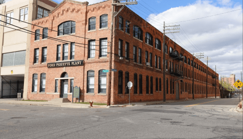 Historic Ford Piquette Plant, a red brick building with large windows, located at a street intersection.
