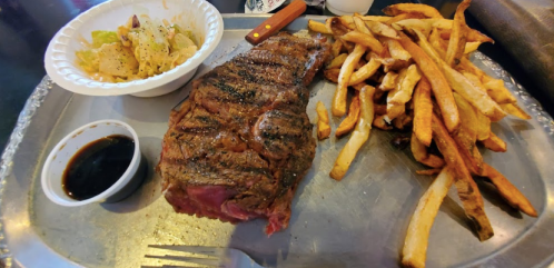 A grilled steak on a metal tray with fries and a side salad, accompanied by a small cup of sauce.
