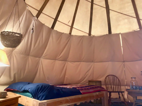 Interior of a cozy yurt with a bed, wooden chair, and soft lighting, featuring fabric walls and rustic decor.