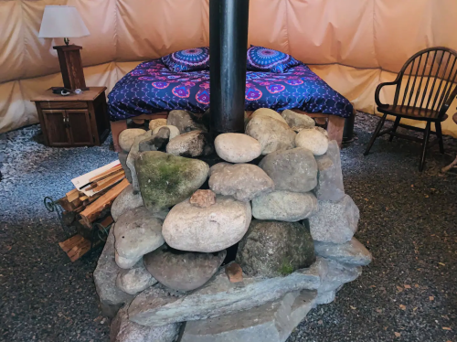 Cozy interior of a yurt featuring a stone fireplace, a bed with colorful bedding, and a wooden chair.
