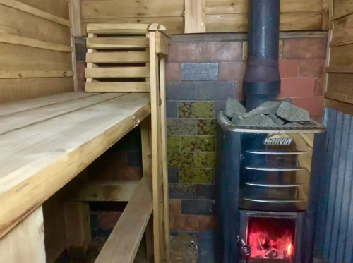 A cozy sauna interior featuring a wooden bench, a stone wall, and a metal stove with glowing embers.