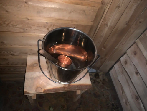 A metal bucket with water and a small cup sits on a wooden stool in a rustic setting.