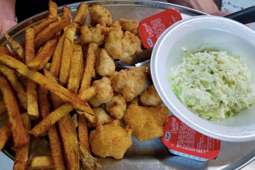 A metal platter with fried fish, shrimp, fries, and coleslaw, accompanied by two cups of cocktail sauce.
