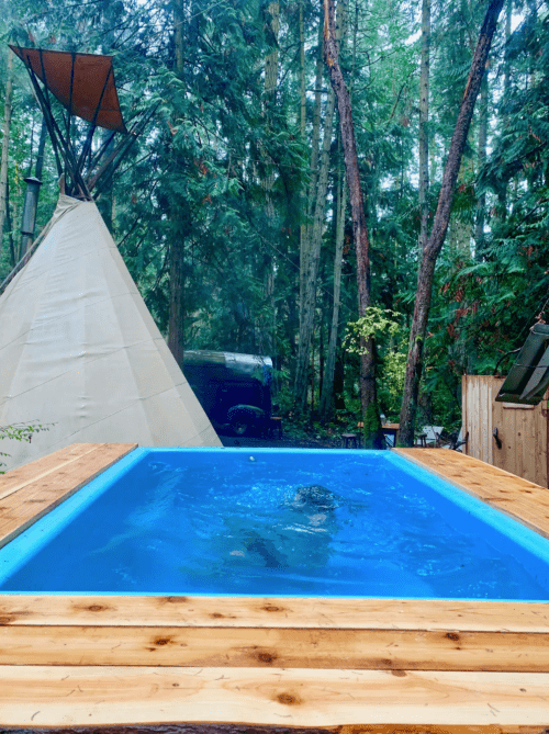 A blue hot tub surrounded by trees, with a teepee and a vehicle in the background.