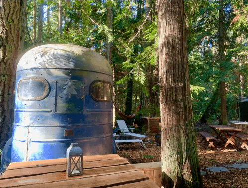 A vintage blue trailer nestled among tall trees in a serene forest setting, with outdoor furniture and a lantern nearby.