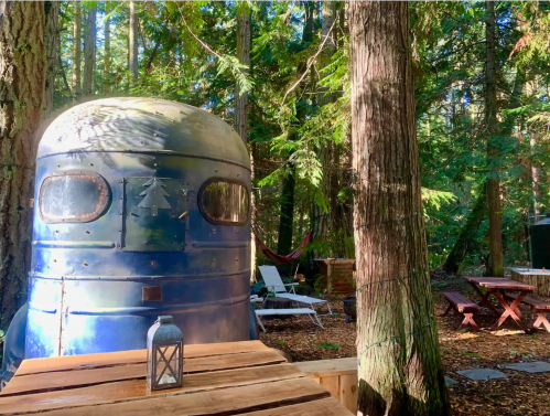 A vintage blue trailer nestled among tall trees in a serene forest setting, with outdoor furniture and a lantern nearby.