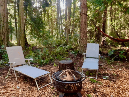 A serene forest campsite with two lounge chairs, a fire pit, and a hammock among tall trees and lush greenery.