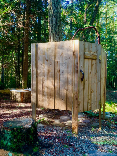 A wooden outdoor shower stall surrounded by trees and gravel, with a rustic design and a small stool nearby.